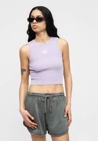 Γυναίκα που φοράει μαύρα ορθογώνια γυαλιά ηλίου, λιλά αμάνικη κορυφή crop top και γκρι σορτς με κορδόνι, στέκεται με το ένα χέρι χαλαρό στο πλευρό της.