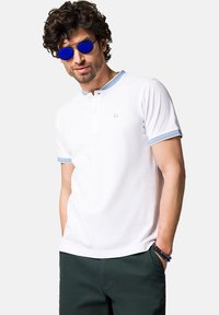 Biała koszulka polo z jasnoniebieskim kołnierzykiem i wykończeniami rękawów, z zapięciem na guziki i małym logo na piersi. Nosi ją w połączeniu z ciemnymi spodniami.