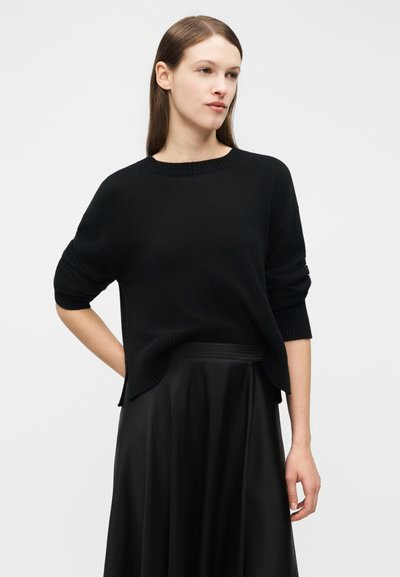 Młoda kobieta z długimi brązowymi włosami nosi luźny czarny sweter i zwiewną czarną spódnicę, stojąc na tle jasnego, jednolitego tła.