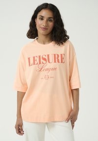 T-shirt surdimensionné couleur pêche, en coton, à manches courtes, avec le texte "LEISURE League" en corail et des détails floraux, coupe décontractée.