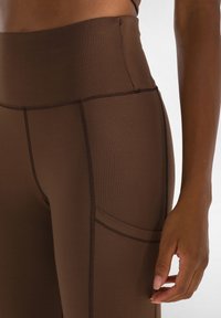 Leggings marroni a vita alta con una texture liscia, dotati di cuciture piatte e una tasca laterale, che offrono una silhouette aderente.