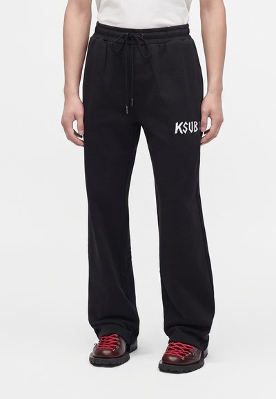 Personne portant un pantalon noir à cordon avec un texte blanc "KSUBI" sur la cuisse droite et des bottes marron foncé avec des lacets rouge vif.