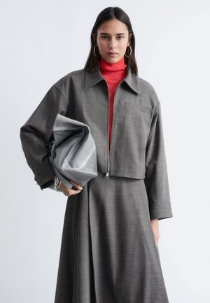 Kvinde iført grå kort jakke og nederdel med rød højhalset trøje, holder stor grå clutchtaske, står mod en ensfarvet lyse baggrund.