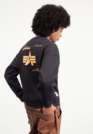 Felpa nera con logo arancione sul retro, polsini e orlo a costine, indossata con pantaloni cargo marroni caratterizzati da dettagli sulle tasche.