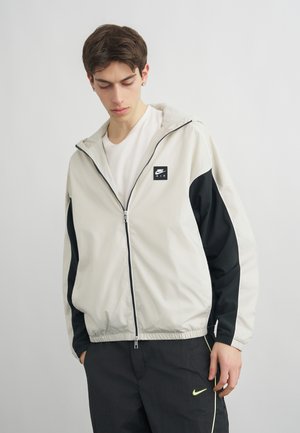 Νεαρός άνδρας που φοράει λευκό και μαύρο φούτερ με φερμουάρ Nike Air και μαύρο παντελόνι Nike με νέον λογότυπο, κοιτάζοντας προς τα κάτω, με απλό γκρι φόντο.