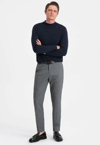 Navy-blauer gerippter Strickpullover, graue strukturierte Stoffhose und schwarze Schnürschuhe. Das Modell steht mit verschänkten Armen vor einem weißen Hintergrund.