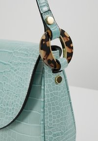 Sac à main bleu clair à motif crocodile avec bouton doré et accent circulaire en écaille attaché à la sangle.