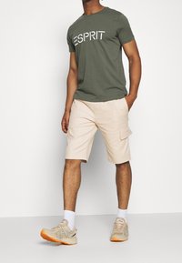 Olivenfarbener Baumwoll-T-Shirt mit weißer "ESPRIT"-Schriftzug; beigen Cargo-Shorts mit Taschen; hellbeige Sportschuhe mit orangefarbener Akzenten.