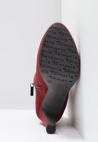 Red suede high-heeled shoe with a pointed toe, side zipper detail, and a textured black rubber sole featuring repeated branding. Size 37.