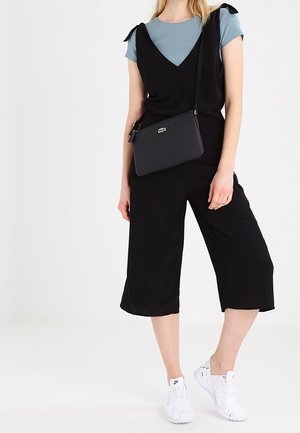 Combinaison sans manches noire avec des jambes larges et bouffantes, associée à un t-shirt bleu clair et un sac bandoulière noir. Des baskets blanches complètent la tenue.