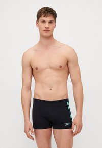 Černé plavky vyrobené z elastického materiálu, s postranním panelem se zeleným vzorem a logem Speedo na dolní části předního dílu.