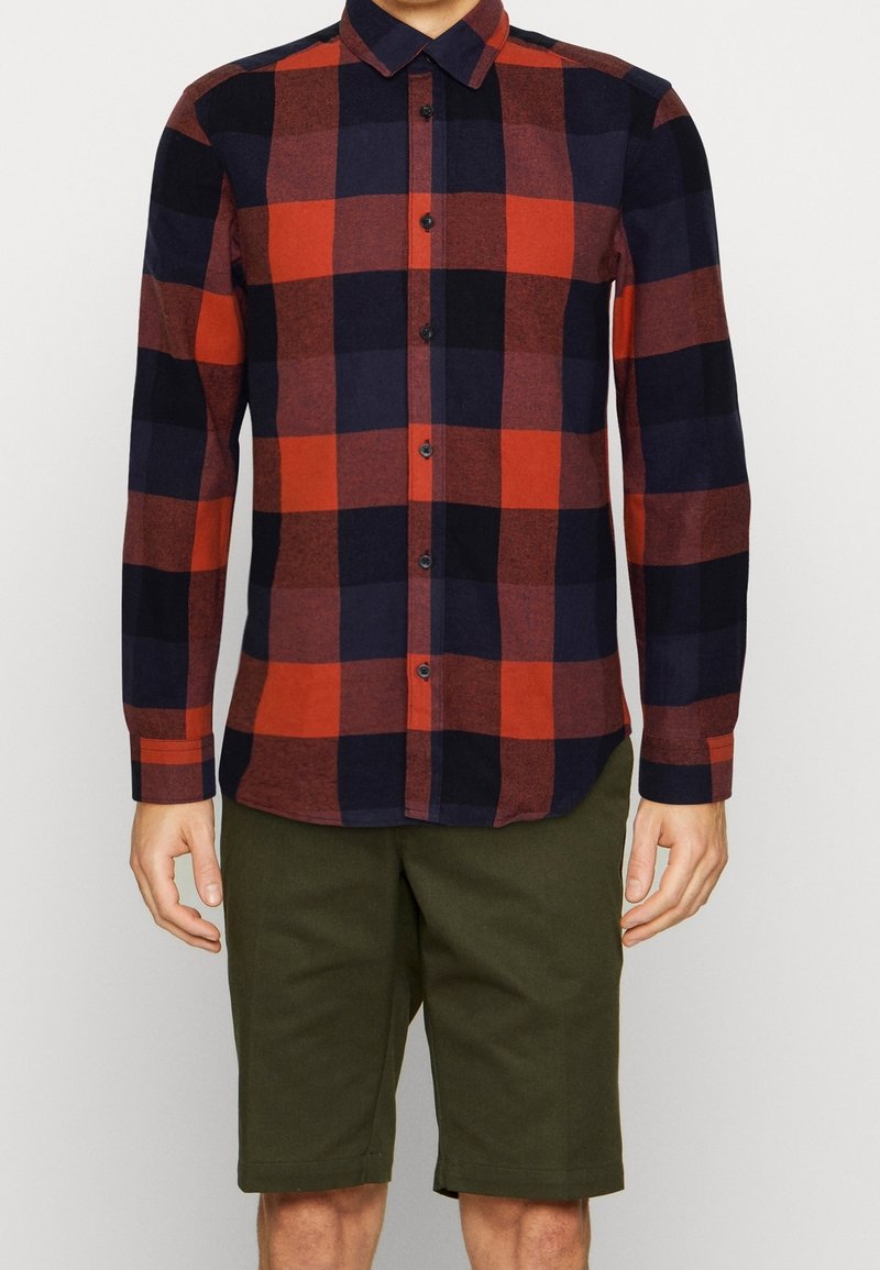 Hombre vestido con una camisa de cuadros roja y negra de manga larga y pantalones cortos oscuros de hasta la rodilla, de pie frente a un fondo sencillo.