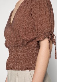 Brun blus med prickar och djupt V-hals, smockad midja och korta puffärmar som är knutna vid ärmsluten. Lättviktsmaterial med texturerad yta.