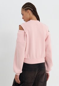 Rosa verkürzter Kapuzenpullover mit offenem Schulterdesign, gerippten Bündchen und Saum. Tragen mit dunklen Cargo-Hosen, die große Taschen haben.