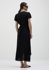 Donna che indossa un abito midi nero con maniche a sbuffo corte e vita elastica, sandali neri e occhiali da sole, in piedi su un pavimento bianco.