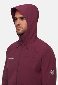 Un homme portant une veste à capuche bordeaux avec le logo Mammut sur la poitrine et le logo de mammouth sur la manche, faisant face à gauche contre un fond uni.