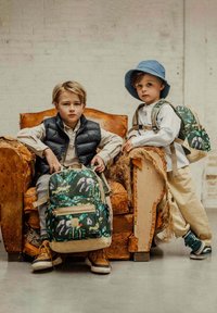 Twee kinderen zitten in een vintage fauteuil. Het kind aan de linkerkant draagt een zwart puffervest en houdt een groene rugzak met dierenprints vast. Het andere kind draagt een licht shirt, beige broek en een blauwe hoed, en draagt een kleinere groene rugzak.
