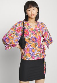 Top envolvente floral em flores vibrantes de rosa, roxo e laranja sobre fundo branco, com mangas largas e cintura atada, combinado com uma saia preta.