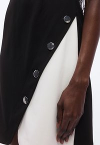 Vestido preto e branco com divisão diagonal, apresentando botões prateados polidos ao longo da costura. O tecido parece liso com um design estruturado.