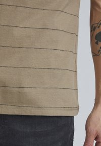Hele pruun T-särk mustade horisontaalsete triipudega, valmistatud pehmest kangast. Omab ümmargust kaelust ja lühikesi varrukaid. All on näha mustad teksad.