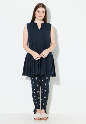 Granatowy top bez rękawów z plisowanym wzorem, w połączeniu z granatowymi legginsami z geometrycznym wzorem w kolorze kremowym i beżowym. Beżowe sandały.