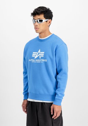Lichtblauwe sweatshirt met een ronde hals, ribbelboorden en -zoom. Bevat een groot wit logo op de borst. Draagt met zwarte shorts.