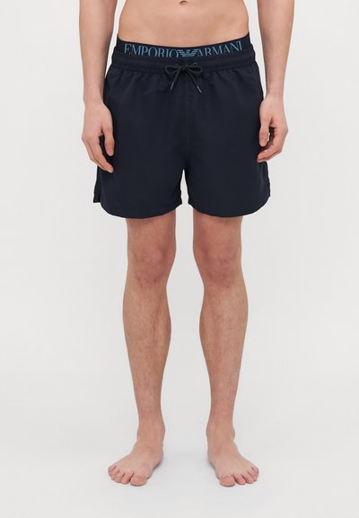 Shorts de bain bleu marine avec taille élastique, cordon de serrage et poches latérales. Tissu lisse, design minimaliste sans motifs ni accents.