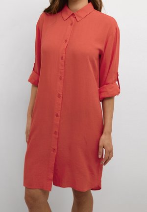 Kvinde iført en løstsiddende koralfarvet knap-ned-kjole med opkørte trekvartærmer foran en ensfarvet baggrund.