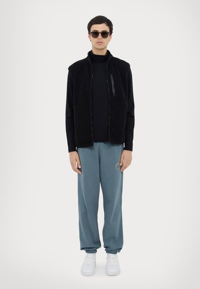 Gilet en polaire noir sur un col roulé noir, associé à un pantalon de survêtement bleu clair avec un motif graphique, et des baskets blanches. Lunettes de soleil portées.