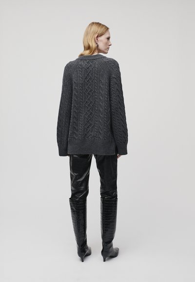 Blonde Frau, die von hinten zu sehen ist, trägt einen dunkelgrauen Zopfmuster-Pullover, schwarze Lederhosen und kniehohe schwarze strukturierten Stiefel.