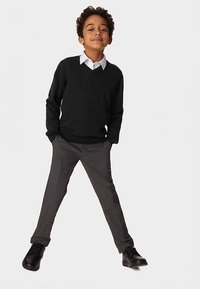 Suéter negro de cuello en V, camisa blanca con cuello, pantalones grises y zapatos negros. Niño posa con los brazos ligeramente abiertos, sonriendo.