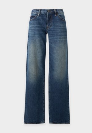 Femme aux longs cheveux foncés portant un haut blanc à manches courtes avec col et un jean bleu à jambes larges avec des chaussures noires pointues, debout les mains dans les poches.