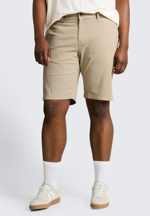 Man draagt beige knielange shorts, witte sportsokken en beige sneakers met witte strepen, staand tegen een effen achtergrond.