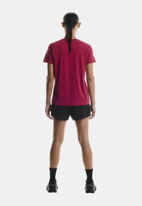 Lahka rdeča športna majica s kratkimi rokavi, kombinirana s črnimi kratkimi hlačami. Ima gladko teksturo in minimalističen dizajn.