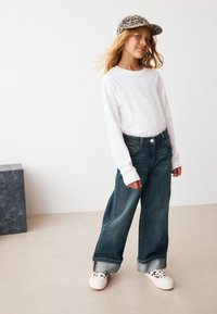 Biela košeľa s dlhým rukávom, široké tmavomodré rifle s ohrnutými lemami a biele tenisky. Čiapka má vzor leopardieho potlače.