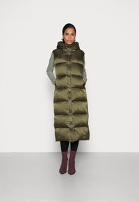 Gilet imbottito verde oliva con cappuccio, con chiusura a zip e design a righe verticali. Indossato sopra una maglietta grigia a maniche lunghe e pantaloni neri.