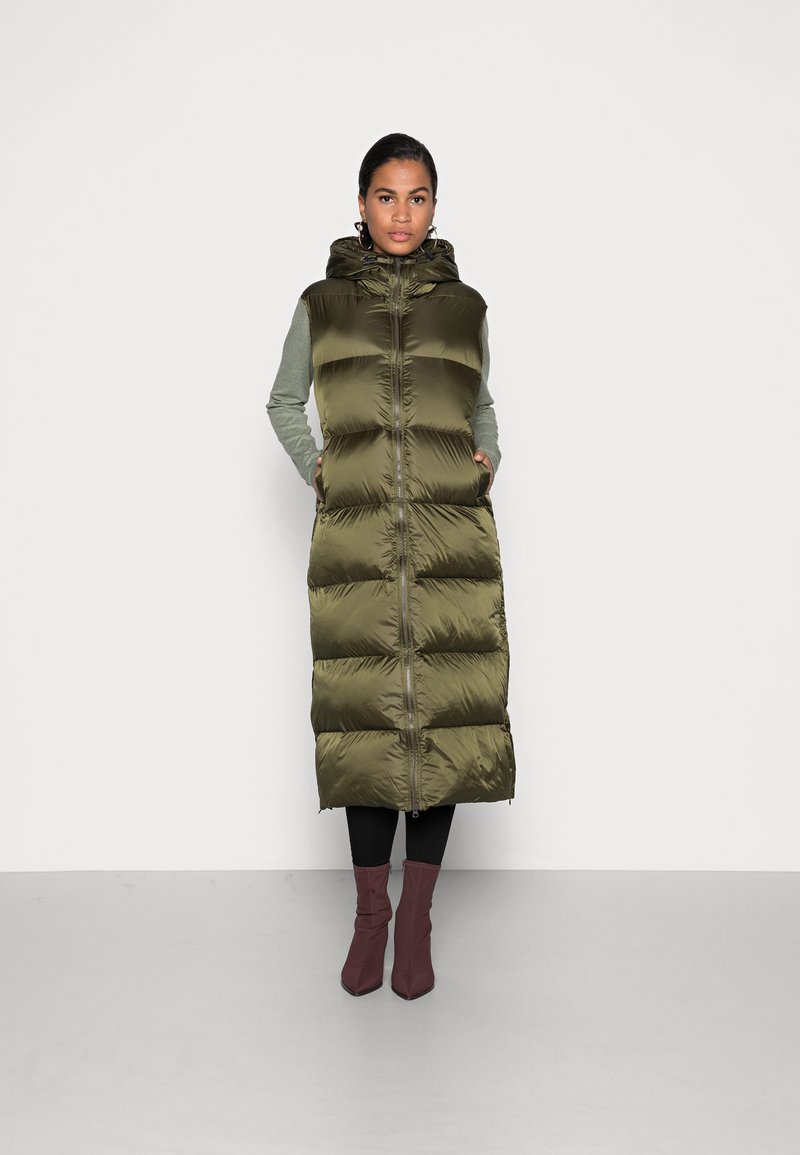 Gilet imbottito verde oliva con cappuccio, con chiusura a zip e design a righe verticali. Indossato sopra una maglietta grigia a maniche lunghe e pantaloni neri.