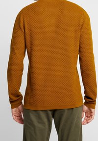 Pull marron texturé avec un motif de tricot gaufré, manches longues et poignets côtelés. Porté avec un pantalon vert olive.