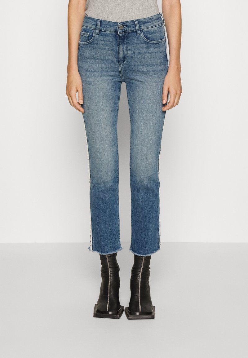 Modré džínové kalhoty s rovnými nohavicemi, roztrhaným lemem a bočními pruhy. Nosené s černými kotníkovými botami se čtveratou špičkou. Hladká textura.