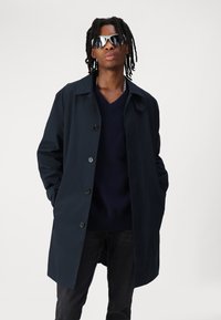 Marineblaue Trenchcoat mit Reverskragen, Knopfverschluss und seitlichen Taschen. Getragen über einem dunklen Strickpullover und schwarzen Jeans.