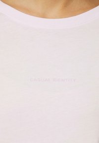 T-shirt rose clair avec un col rond et une texture douce, arborant le texte "IDENTITÉ CASUAL" en lettrage discret et légèrement en relief.
