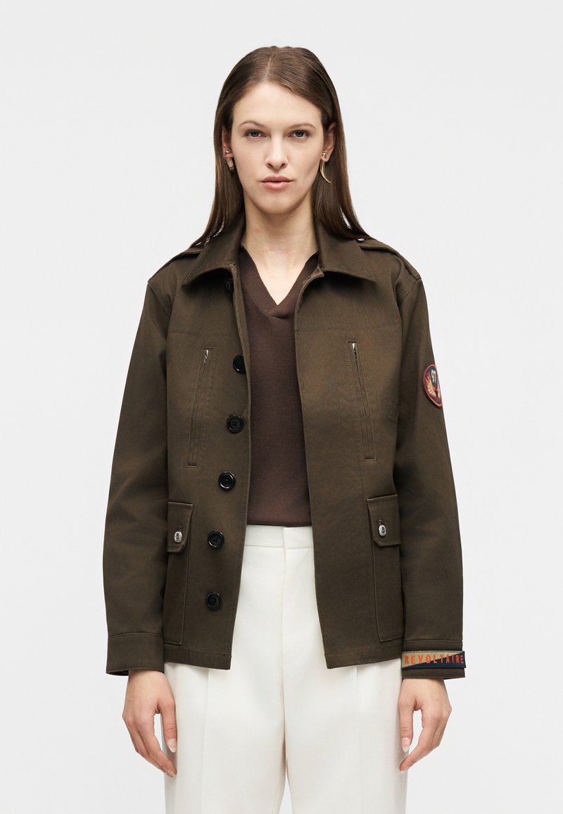 Γυναίκα που φοράει σκούρο καφέ σακ άκι με κουμπιά και επιθεματικό στο αριστερό μανίκι και άσπρο παντελόνι, στέκεται μπροστά σε ένα απλό ανοιχτόχρωμο φόντο.