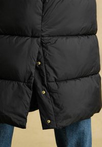 Černá péřová bunda se čtvercovým prošíváním, lesklou texturou, zlatými patentkami a rovnoměrným lemem. Kombinována s džíny.