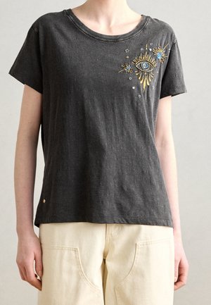 Osoba ubrana w ciemnoszary T-shirt z haftowanymi niebiesko-złotymi wzorami oka i gwiazdy na górnej lewej części piersi, w połączeniu z jasno beżowymi spodniami.