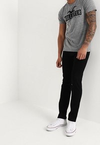 Camiseta gris de manga corta con logotipo negro, combinada con jeans negros y zapatillas blancas. El conjunto presenta una silueta casual y ajustada.
