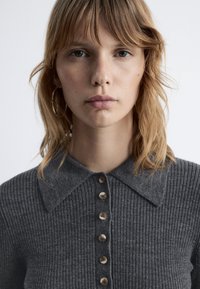 Grauer gerippter Pullover mit Kragen, der eine Knopfreihe auf der Vorderseite und eine weiche Textur aufweist. Detaillierte Nahaufnahme.