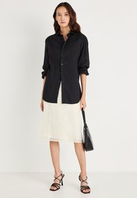 Camisa preta de botão com mangas longas, sobreposta a uma saia branca clara, bordada. Modelo segura uma mala preta. Sapatos de salto pretos com tiras completam o visual.