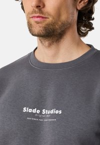 Felpa grigia con scollatura a girocollo. Presenta testo stampato bianco: "Slade Studios Original Wear" e "DESIGNATO PER LA TUA LIBERTÀ". Design pulito.