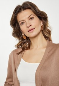 Cardigan tricotat de culoare maro peste un top alb, având un design în formă de V. Păr ondulat și cercei circulari cu un amestec de culori și texturi.