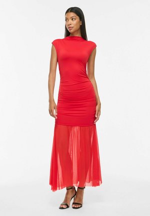 Woman with long dark hair wearing a fitted red dress with a sheer lower skirt and black high-heeled sandals, standing against a white background.